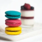 Macaron berwarna-warni tersusun di atas piring dengan dessert buah beri di latar belakang